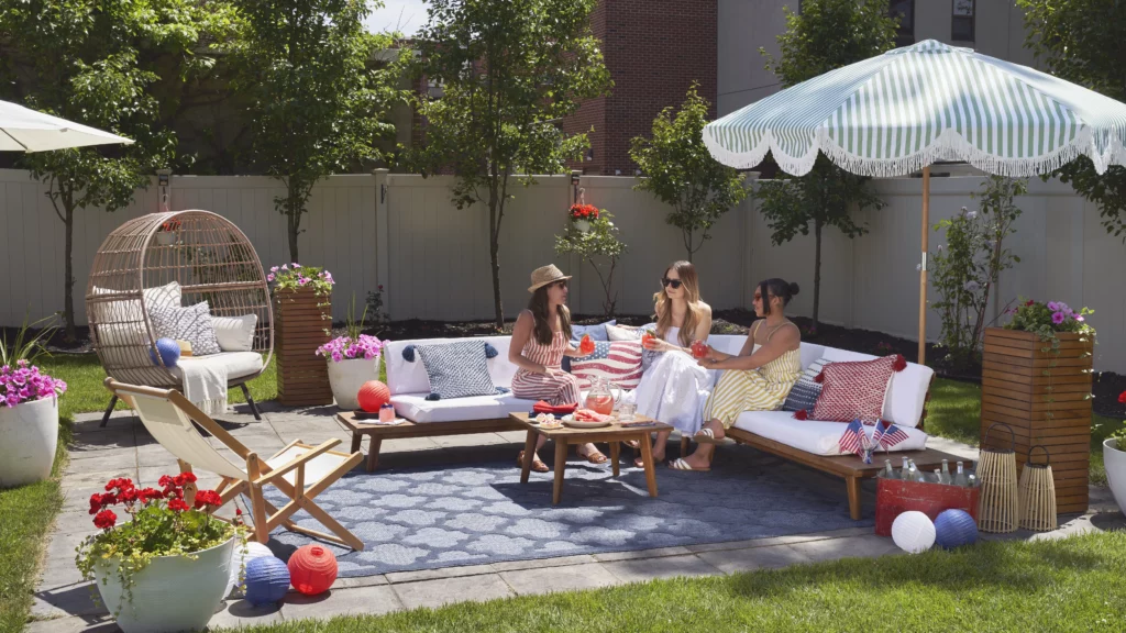Three women sitting on outdoor patio furniture around a blue indoor/outdoor area rug on a stone patio, surrounded by colorful flowers and summer decor.