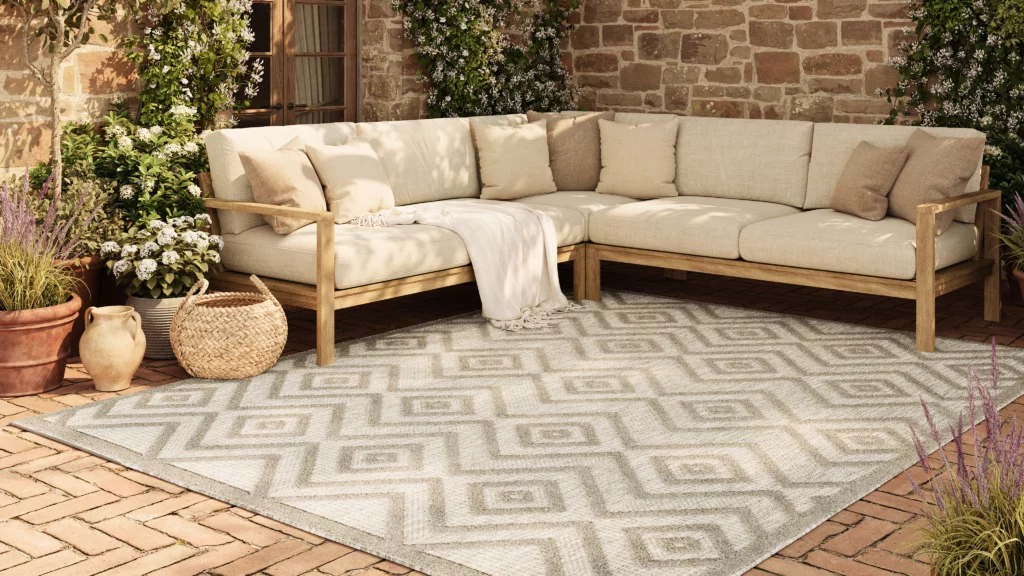 Nourison Home Versatile NRV01 silver grey geometric outdoor rug on a brick patio with teak sectional sofa and flowering potted plants against a stone wall