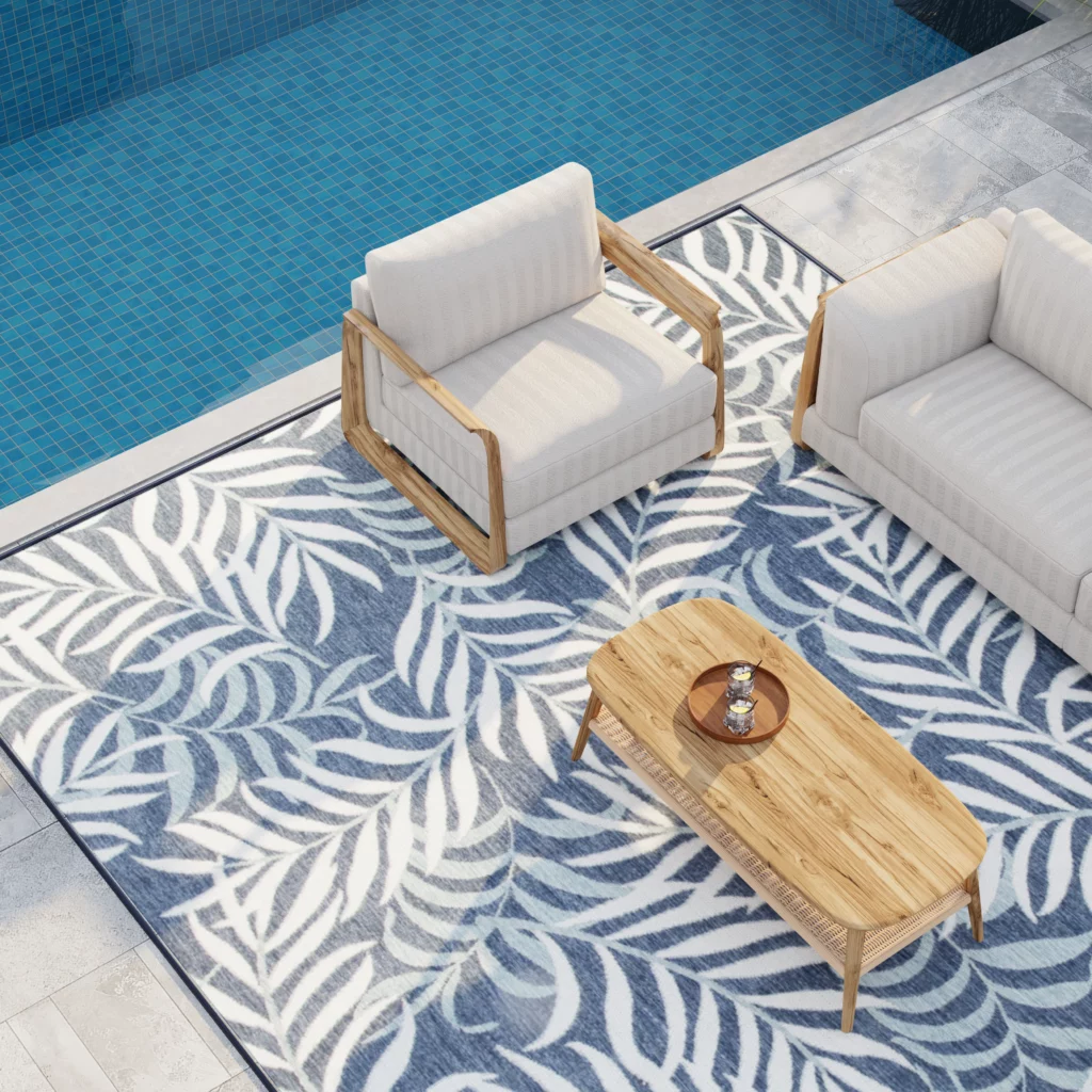 Overhead view of a navy blue botanical indoor/outdoor area rug under teak armchairs and a coffee table at a poolside patio.