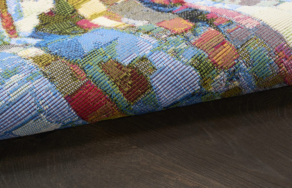 Close-up of a multicolor flatwoven outdoor rug rolled on a wood surface, showing the open weave construction and backing detail.