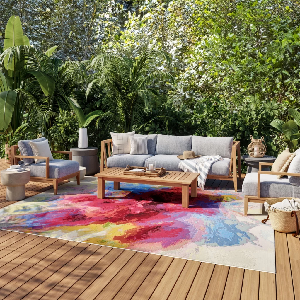Ivory and multicolor abstract watercolor indoor/outdoor area rug on a wood deck patio with teak sofa and armchairs surrounded by tropical greenery.