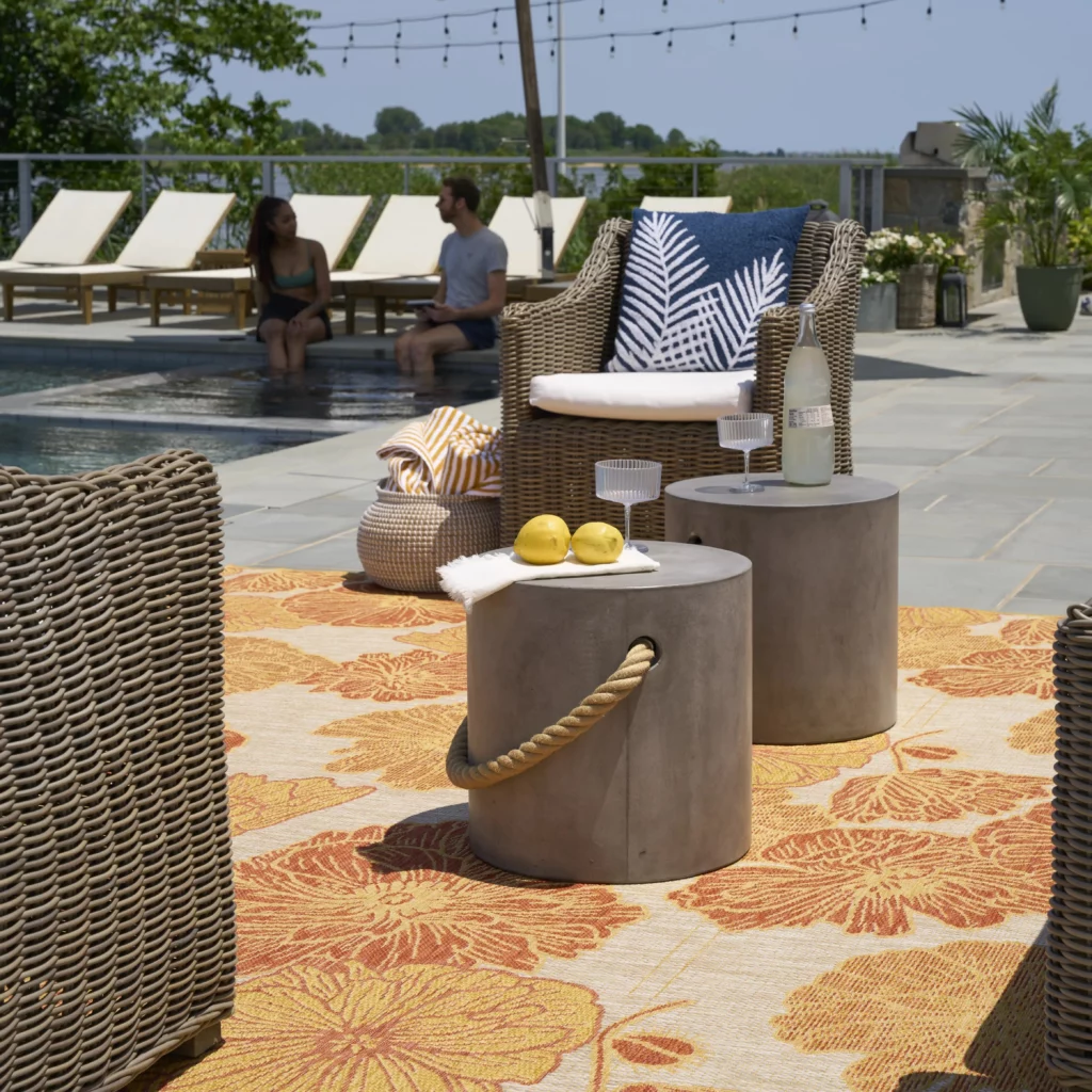 Coral floral indoor/outdoor area rug under wicker chairs and concrete side tables on a stone pool patio, with two people seated by the water in the background.
