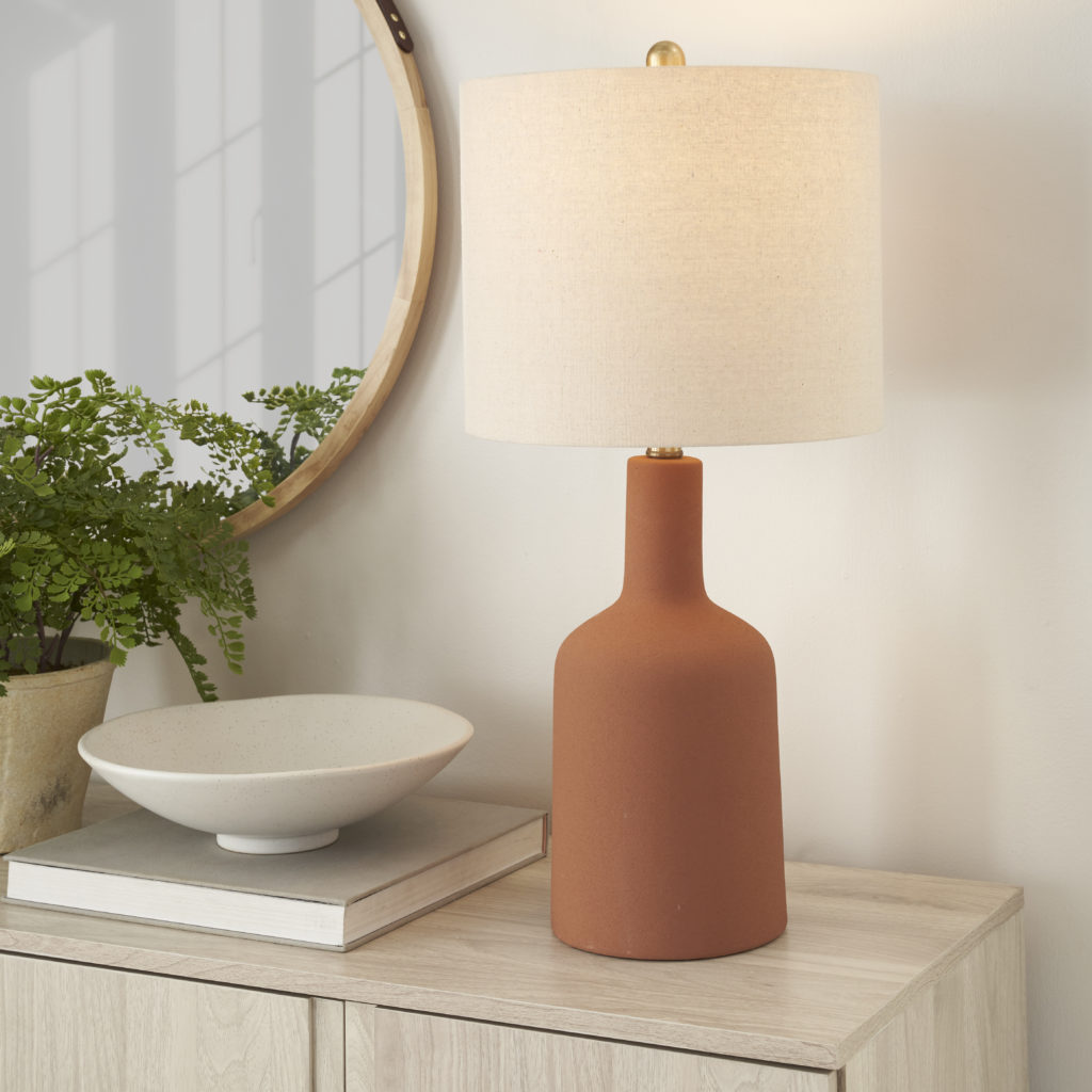 Terracotta ceramic table lamp with a linen drum shade on a light wood console, styled with a round mirror, potted fern, and white ceramic bowl