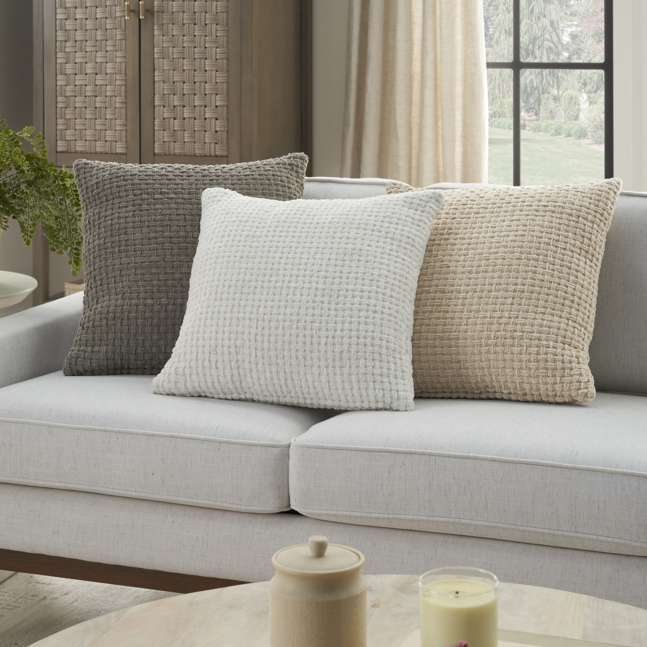 Three textured woven throw pillows in beige, white, and gray displayed on a light gray sofa in a neutral living room
