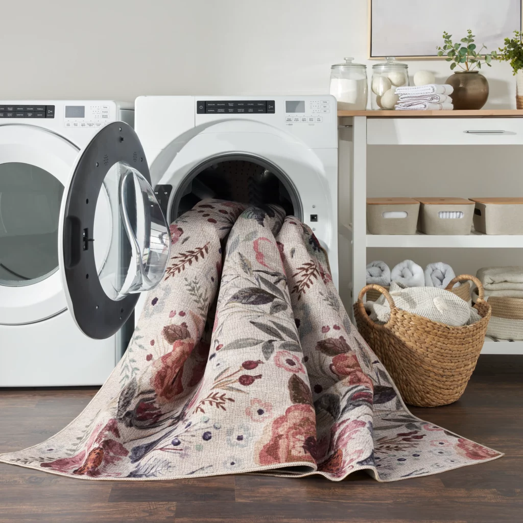 Nourison Washables NWB01 Ivory Pink 2x3 floral area rug being loaded into a front-load washing machine in a clean, organized laundry room with folded towels and woven baskets