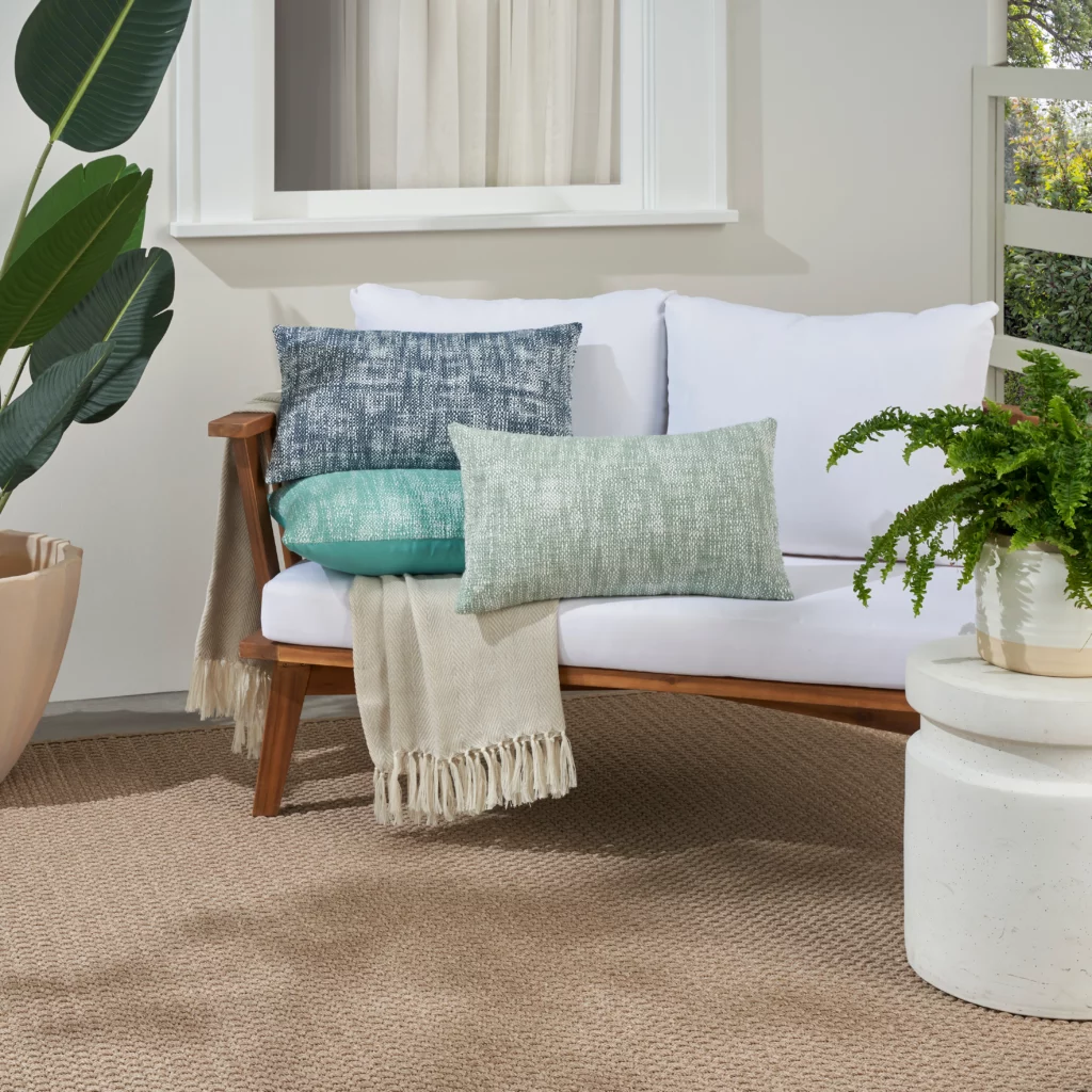 Mina Victory aqua 12 by 21 lumbar outdoor pillow on a neutral wood-frame bench with a light-colored outdoor rug and potted plants in the background