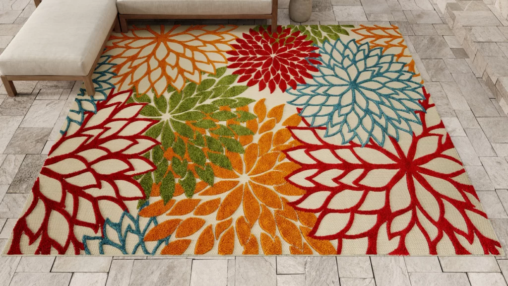Nourison Aloha ALH05 outdoor rug in green with a large-scale multicolor dahlia floral pattern on an ivory ground, styled on a stone tile patio with a wood-frame ottoman