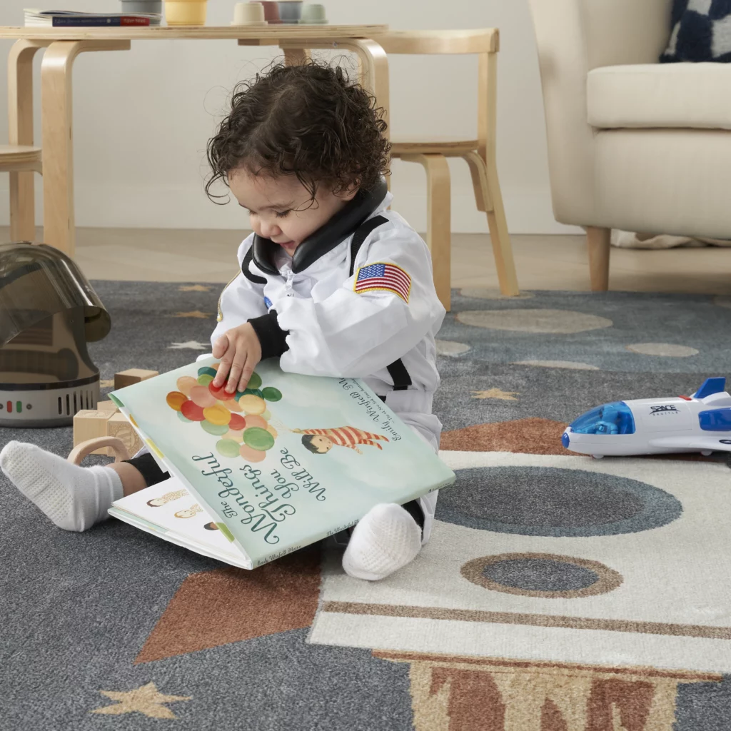 Toddler in an astronaut costume sitting on the Nourison Astra ASW06 Sky Blue 7x9 area rug reading a picture book, surrounded by space toys and wooden blocks in a playroom