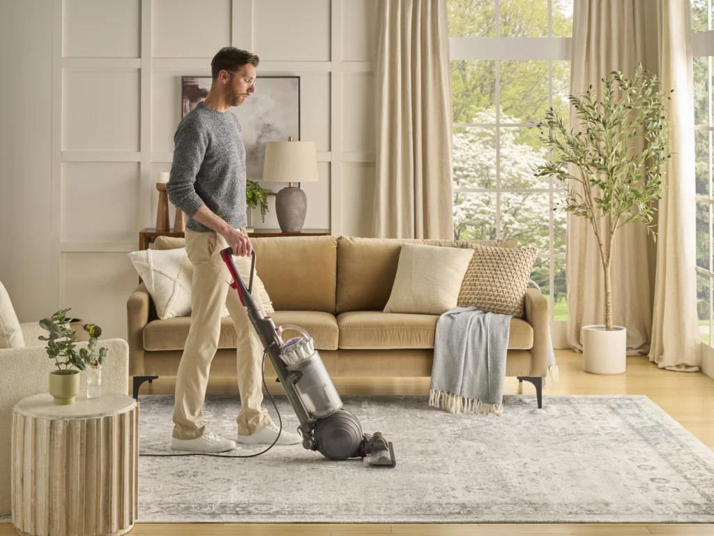 Man vacuuming a light gray and ivory distressed traditional area rug in a warm, neutral living room with a tan sofa and large windows