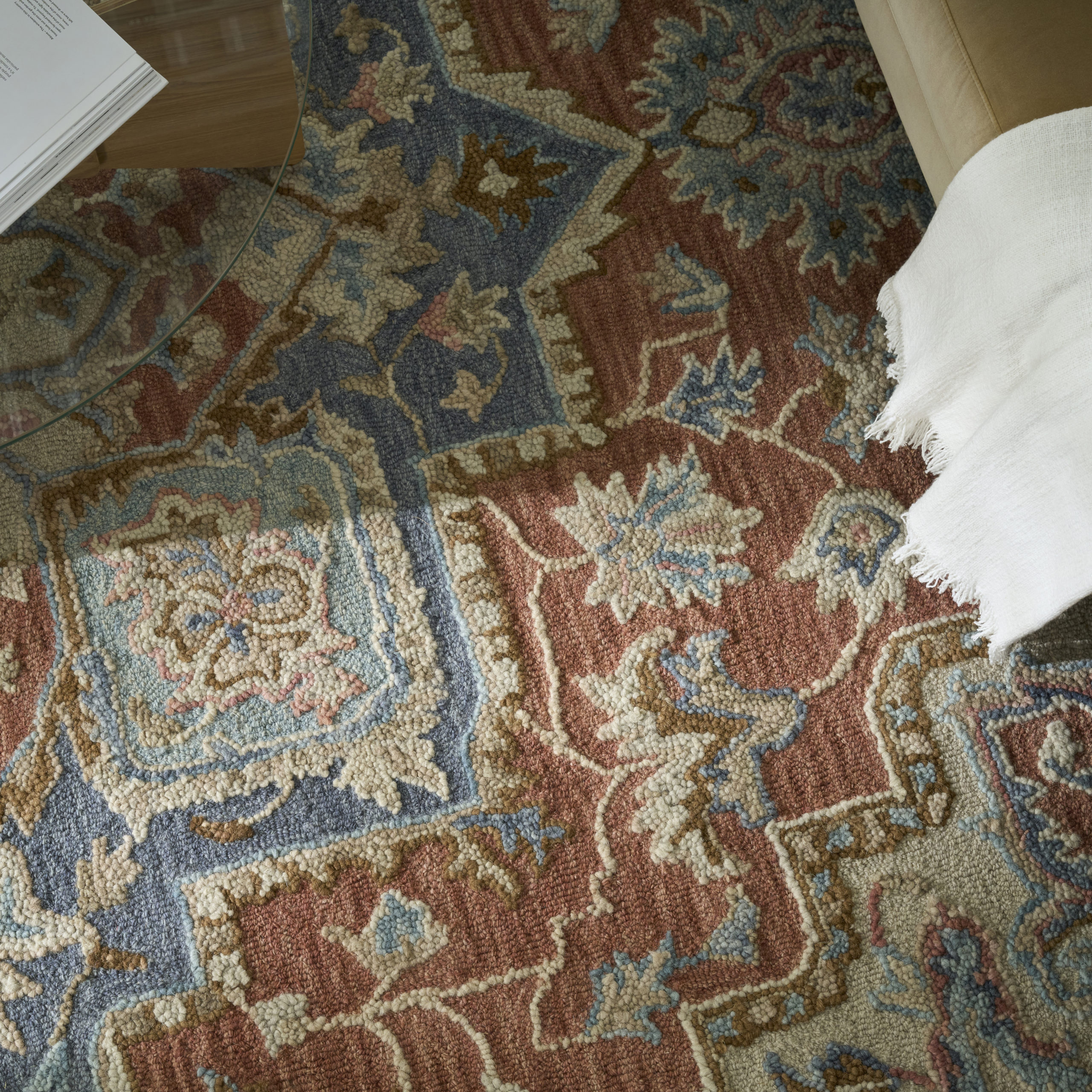 Close-up overhead view of a hand-tufted wool area rug in rust and blue with a traditional Persian floral pattern, with a white fringe throw partially visible