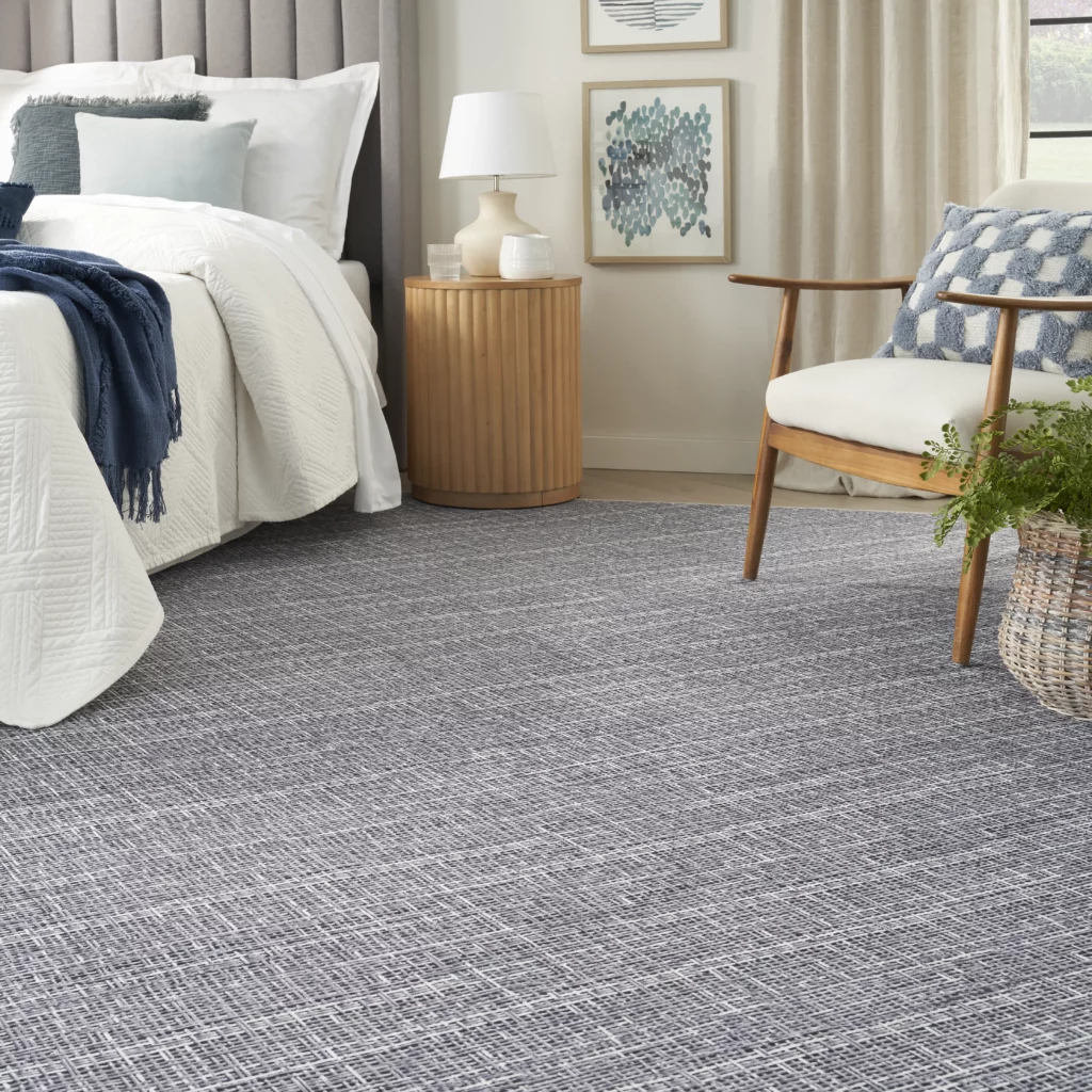 Nourison Washables NWB08 Grey 8x10 textured solid area rug in a calm bedroom with a white quilt, navy throw, fluted wood nightstand, and a mid-century accent chair