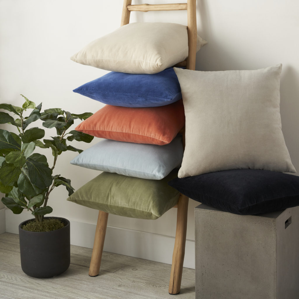 Mina Victory Sofia velvet throw pillows stacked on a wooden ladder in assorted colors including coral, navy, sage, and ivory, with a potted plant nearby