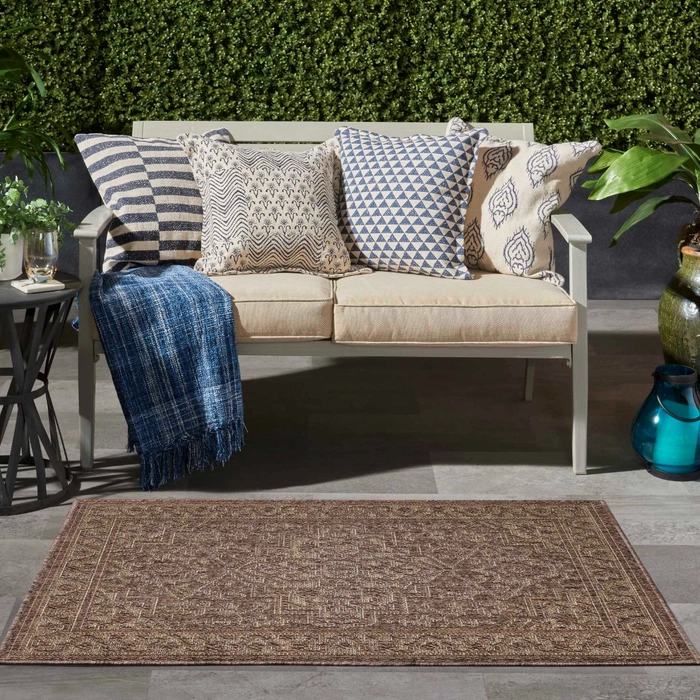 LM05 brown indoor outdoor flatweave area rug on a patio in front of a gray wood bench with patterned throw pillows and greenery