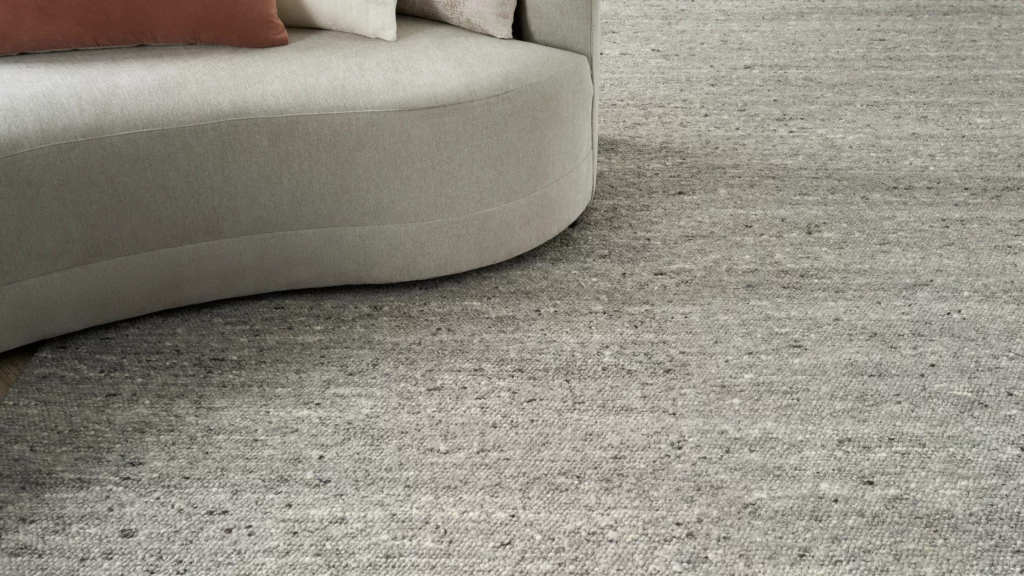 Close-up of a grey wool area rug with a textured looped pile next to the base of a curved light grey sofa