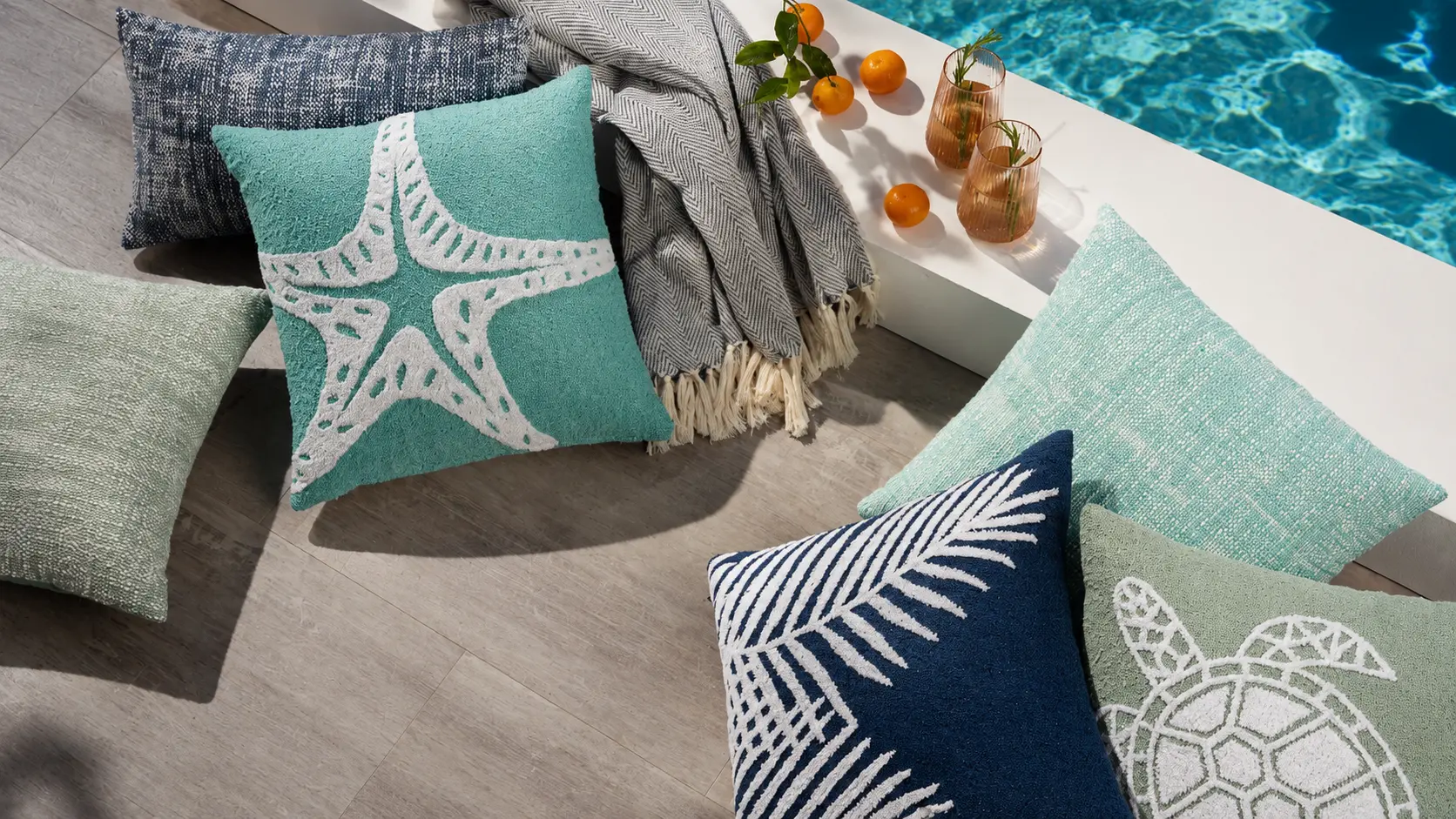 Assortment of Nourison Mina Victory outdoor pillows with coastal motifs including a starfish, sea turtle, and palm frond in teal, navy, and sage green, arranged beside a swimming pool