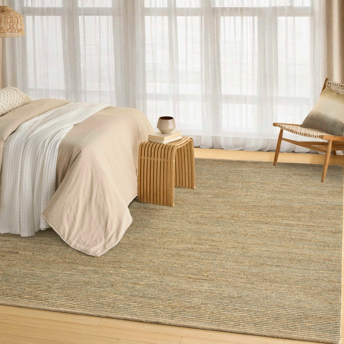 Dusty sage textured area rug with a bordered design styled in a serene bedroom with a linen bed, slatted wood nightstand, woven accent chair, and sheer white curtains