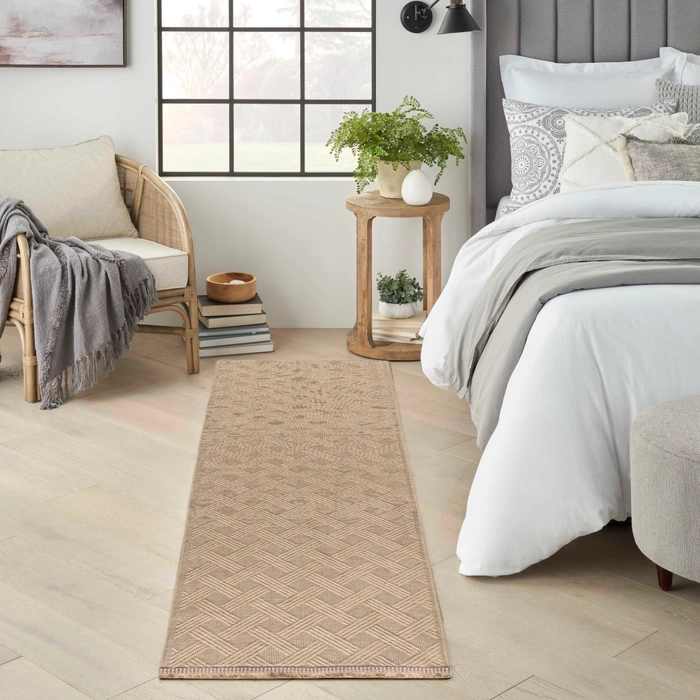 Natural toned geometric patterned washable runner rug styled alongside a bed in a cozy bedroom with light hardwood floors, a rattan accent chair with a grey throw, and a round wood nightstand with plants