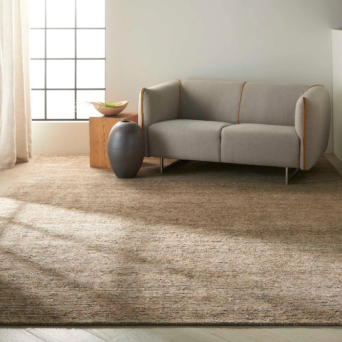Amber toned solid textured area rug styled in a minimalist living room with a grey loveseat with mustard piping, a raw wood side table, and a dark ceramic vase