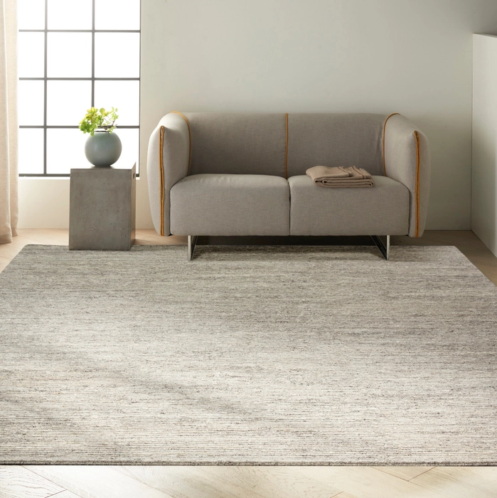 Grey variegated solid area rug styled in a minimalist living room with a rounded grey loveseat with mustard piping, a concrete pedestal side table, and a sage ceramic vase