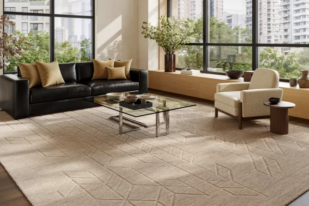 Nourison Home Alessia ALE01 Beige wool area rug with a carved geometric pattern beneath a glass coffee table in a sunlit living room with a black leather sofa.
